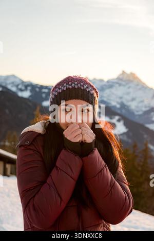 Eine Frau im dicken Wintermantel steht in einer verschneiten Landschaft in der Schweiz, mit Sonnenlicht, das schneebedeckte Gipfel beleuchtet und die Schönheit eines sonnigen Ko einfängt Stockfoto