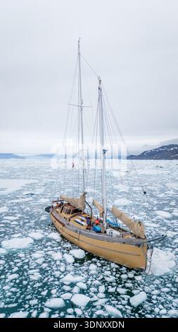 Ein Segelboot navigiert anmutig durch die eisigen Gewässer der Davis-Straße in Grönland, umgeben von schwimmenden Eisbergen und weitläufigen Meereslandschaften unter einem Clo Stockfoto