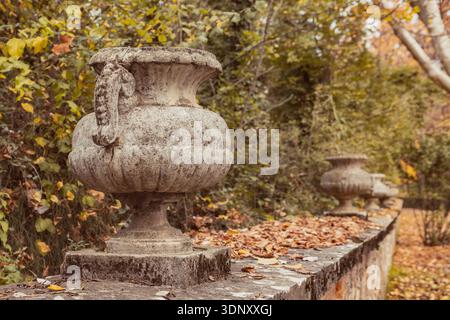 Ein Steinzaun mit verzierten Vasen und einer Hecke, der an einen Pfad grenzt, der mit gelben und orangen Herbstblättern bedeckt ist. Die heitere Schönheit des Herbstes, die Natur, die s Stockfoto