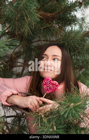 Eine Frau in rosa Jacke hält einen rosa Lutscher in Herzform und lehnt sich spielerisch an die grünen Zweige eines Weihnachtsbaums. Ihr fröhlicher Ausdruck Stockfoto