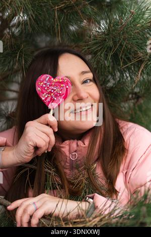 Eine fröhliche Frau in rosa Jacke hält spielerisch einen Herzlutscher nahe ihrem Auge, eingerahmt von den Zweigen eines Weihnachtsbaums, um das fest festzuhalten Stockfoto