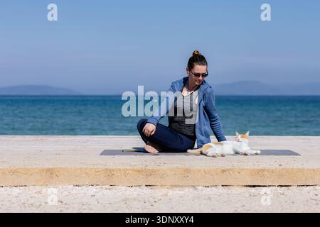 Eine Frau übt Yoga auf einer Matte am Meer und sitzt in einer entspannten Position, während sich eine Katze in der Nähe ausdehnt. Die friedliche Szene zeigt Harmonie und Rela Stockfoto