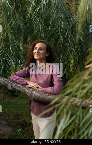 Frau in lässiger Kleidung mit lockigen Haaren lehnt sich auf einen Baumzweig in einer ruhigen Parklandschaft, umgeben von üppigem Grün, die Entspannung und Kontemp zum Ausdruck bringt Stockfoto