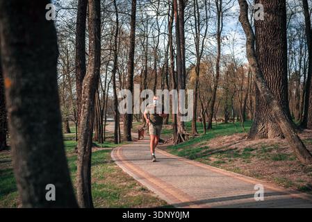 Ein junger Mann joggt auf einem malerischen Pfad in einem üppigen Park, umgeben von hohen Bäumen. Er konzentriert sich auf seine Fitness-Routine und genießt die frische Outdoor A Stockfoto