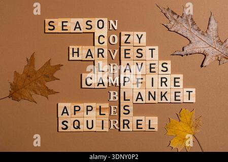 Ein kreatives Kreuzworträtsel aus Holzblöcken, die auf braunem Hintergrund angeordnet sind und mit Herbstblättern verziert sind. Die Worte erinnern an einen gemütlichen, saisonalen geldautomaten Stockfoto