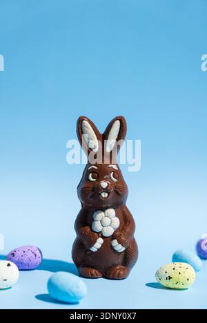 Ein Schokoladen-Osterhase mit komplizierten Details steht auf einem blauen Hintergrund, umgeben von bunten gesprenkelten Eiern. Es ist ein kreatives Stillleben Stockfoto