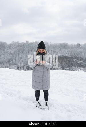 Eine Frau steht draußen in einer verschneiten Winterlandschaft, warm gekleidet mit Mantel und Hut, hält ein heißes Getränk in der Hand und genießt den Lebensstil bei kaltem Wetter inmitten Stockfoto