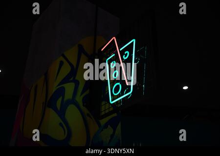 Künstlerisches Neonschild mit geometrischem Design in Cyan und Pink vor dunklem Hintergrund mit gelben Graffiti, die nachts in Breslau einfangen Stockfoto