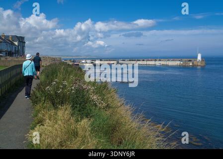 Seahouses Harbour. Blauer Himmel und Sonnenschein. Blühende Pflanzen und Coastal Path Walkers. Seahouses ist ein beliebtes Fischerdorf am Meer in Northumberland. Stockfoto