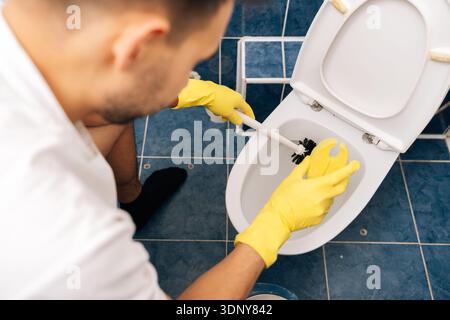 Hochwinkelansicht des Mannes in gelben Schutzhandschuhen, die Toilettenschüssel mit Bürste reinigen, die notwendige Hygienewartung und den täglichen Haushalt durchführen Stockfoto