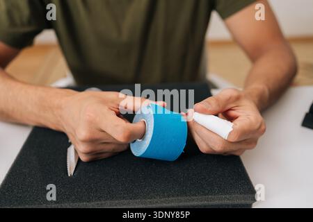 Nahaufnahme eines Mannes, der eine Rolle Kinesiologieband auf dem Tisch hält, um sich auf Muskel-, Gelenk- oder Sehnenschmerzlinderung vorzubereiten und sich selbst zu repräsentieren Stockfoto