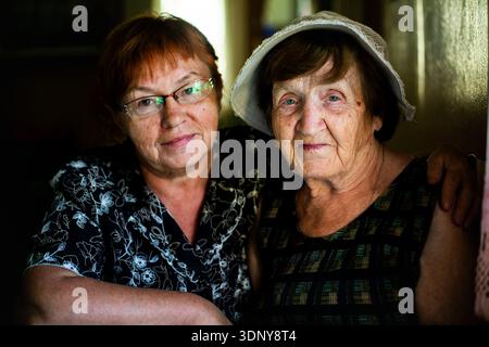 Porträt von zwei Frauen im Haus, einer älteren Mutter und ihrer erwachsenen Tochter, die die Kamera umarmen und betrachten. Weiches natürliches Licht hebt ihre Gesichter hervor Stockfoto