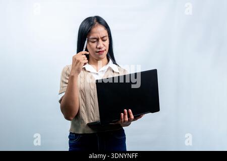 Porträt einer jungen südostasiatischen Frau, die einen Laptop mit einem verwirrten Ausdruck ansieht und einen Stift am Kopf hält. Begriff der Arbeitsbelastung oder des Studiums. Stockfoto