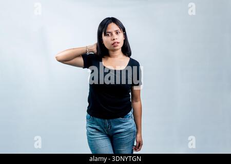 Eine junge Frau in einem schwarzen T-Shirt und einer blauen Jeans posiert selbstbewusst mit einer Hand hinter dem Kopf vor einem sauberen weißen Hintergrund. Stockfoto