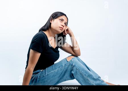 Seitenansicht einer nachdenklichen jungen Frau, die auf einem weißen Boden sitzt, ihren Kopf auf die Hand lehnt und nachdenklich vor einem weißen Hintergrund aufblickt. Stockfoto