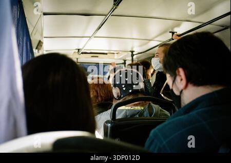 Menschen, die von hinten gesehen werden, sitzen in öffentlichen Verkehrsmitteln, in der Mitte eine Person mit Kappe und Kopfhörern, es gibt Menschen, die Masken auf 35-mm-Film tragen Stockfoto