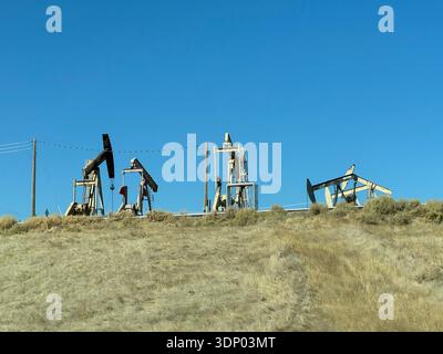 Ölpumpen fördern Rohöl auf einem trockenen, grasbewachsenen Hügel unter klarem blauen Himmel, Maschinen für die Erdölindustrie in einer ländlichen Landschaft. Stockfoto