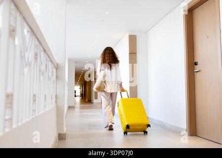 Rückansicht einer erwachsenen Frau, die mit gelbem Koffer durch den Hotelflur ging Stockfoto