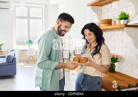 Fröhliches Paar, das Croissant und Kaffee am entspannten Morgen in der Küche teilt Stockfoto