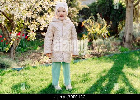 Lächelndes Kleinkind, das in einem blühenden Frühlingsgarten steht, Sonnenlicht und lebhaftes Grün sorgen für einen fröhlichen Familienmoment. Stockfoto