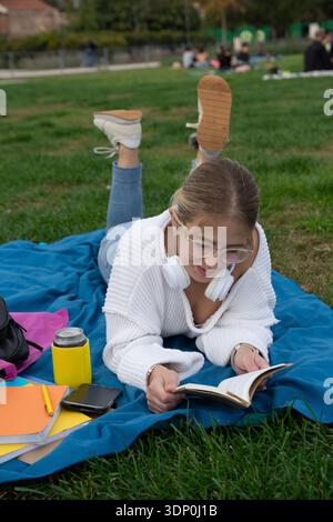 Junge Studentin, die auf blauer Decke auf dem Universitätsrasen liegt, im Freien mit Buch, Kopfhörern, Rucksack und Getränk studiert, entspannt und konzentriert in Geilheit Stockfoto