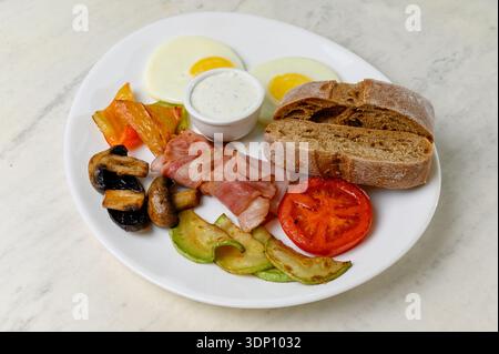 Englisches Frühstück mit Spiegeleiern, gebratenem Speck, Pilzen und Gemüse. Ein Teller mit Spiegeleiern, Speckscheiben, Zucchini-Pilzen und Vollkornbrot Stockfoto