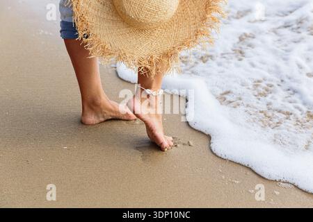 Frauen barfuß Beine und Strohhut in der Nähe von schaumigem Strandwasser Stockfoto