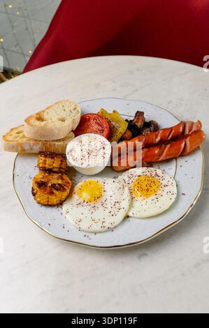 Spiegeleier mit gegrillten Würstchen, Mais und weißer Sauce auf einem Teller. Ein herzhaftes Frühstück mit zwei Eiern, Würstchen, Tomaten und Brotscheiben Stockfoto