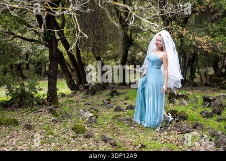 Lächelnde rothaarige Frau in blauem Kleid und Hochzeitsschleier, die im Wald spaziert. Stockfoto
