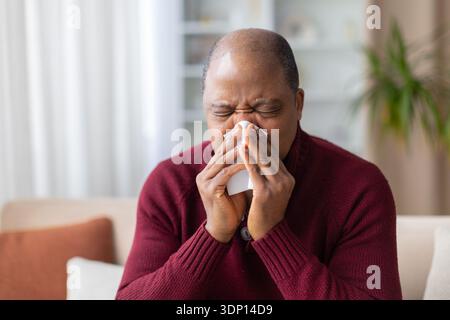Reifer schwarzer Mann niest in Gewebe, während er zu Hause auf dem Sofa sitzt Stockfoto