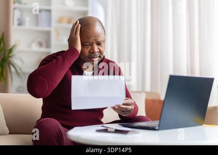 Besorgter schwarzer Mann, der Dokumente hält und den Kopf berührt, während er am Laptop sitzt Stockfoto