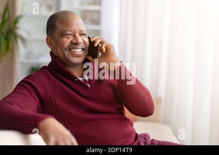 Reifer schwarzer Mann, der auf dem Smartphone spricht, während er zu Hause auf dem Sofa sitzt Stockfoto
