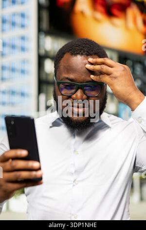 Schwarzer männlicher Erwachsener, der auf seinem Smartphone verstörende Nachrichten erhält und sich besorgt fühlt, frustriert und unzufrieden in der Innenstadt. Erkunden Sie das Stadtzentrum und sein Handy war unzufrieden. Stockfoto