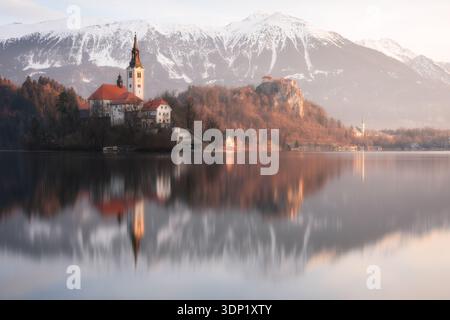 Ein ruhiger Blick auf den Bleder See in Slowenien bei Sonnenuntergang mit der berühmten Kirche der Himmelfahrt auf ihrer kleinen Insel, die sich im ruhigen Wasser spiegelt. Stockfoto