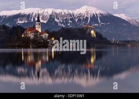Ein ruhiger Blick auf den Bleder See in Slowenien zur blauen Stunde, mit der berühmten Kirche der Himmelfahrt auf ihrer kleinen Insel, die sich im ruhigen Wasser spiegelt. Stockfoto