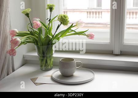Stillleben im Osterfrühling. Tasse Tee, Kaffee und Blumenstrauß auf der Fensterbank. Rosa Tulpen, grüne Nelkenblüten in Vase. Leere Grußkarte Stockfoto