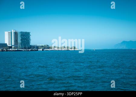 Eine moderne Küstenszene mit einem hohen Glaswohnhaus neben einem Yachthafen mit Segelbooten, ruhigem blauem Meer im Vordergrund. Tróia, Portugal. Stockfoto