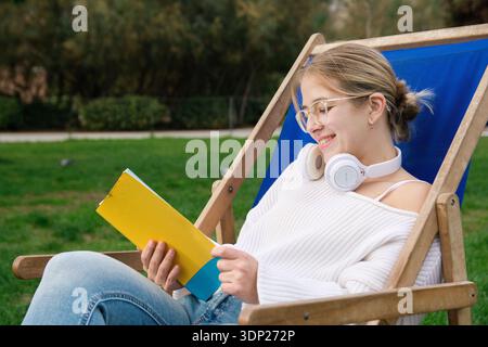 Eine junge Studentin mit Sonnenbrille und Kopfhörern entspannt sich in einem Liegestuhl auf dem Universitätsrasen und lächelt, während sie an einem sonnigen Frühlingstag in ein Buch eingetaucht ist Stockfoto