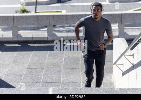 Erwachsener afroamerikanischer Mann, der in Sportbekleidung auf der plaza Treppen läuft, Kopfhörer trägt, Kopierraum Stockfoto