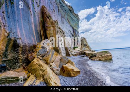 Ein felsiger Strand mit einer Klippe im Hintergrund. Die Klippe ist mit Moos bedeckt und hat ein Loch darin. Der Strand ist mit Felsen und Kieselsteinen bedeckt. Der Himmel ist Stockfoto
