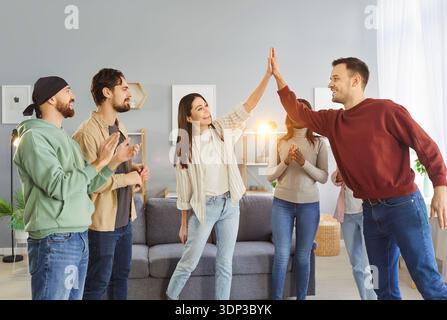 Eine Gruppe verschiedener junger Freunde feiert zu Hause Erfolge, klatscht an den Händen und tauscht High Five aus. Stockfoto