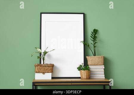 Leerer Rahmen mit Pflanzen und Büchern auf der Bank in der Nähe der grünen Wand Stockfoto