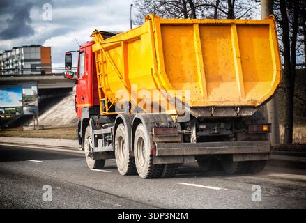 Rückansicht des schweren Muldenkippers, der sich auf asphaltierter Straße bewegt. Das Fahrzeug hat eine rote Kabine und eine große gelbe Ladefläche. Beim cl tritt Staub von den Hinterrädern auf Stockfoto