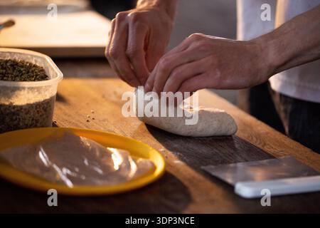 Nahaufnahme der Hände, die mit Teig auf einer rustikalen Holzarbeitsfläche arbeiten. Die Person formt den Teig sorgfältig und bereitet ihn zum Backen vor. Behälter mit getrocknetem Inhalt Stockfoto