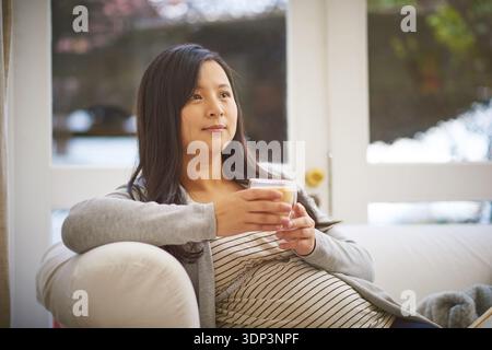 Zuhause, denkende und schwangere Frau auf Sofa, trinken und entspannen mit Tagträumen, Ideen und Erholung. Wohnung, Schwangerschaft und asiatische Person auf Couch, Kräuter Stockfoto