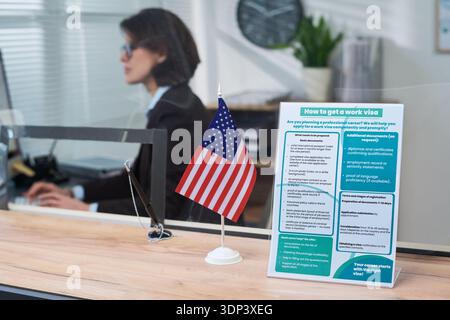 Kaukasische Frau arbeitet am Visumschalter mit Computer, amerikanischer Flagge und Informationsbroschüre für Arbeitsvisum, die auf der Theke im Vordergrund steht, Büroumgebung im Hintergrund sichtbar Stockfoto