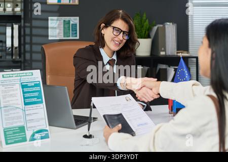 Kaukasische junge Erwachsene Frau mit Brille am Schreibtisch im Visumzentrum und schüttelte während der Konsultation die Hand mit der Bewerberin lateinischer junger erwachsener Frau, die Pass und Dokumente hält Stockfoto