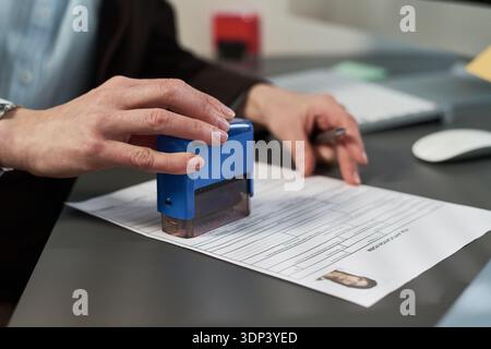 Unbekannte Frau mittleren Alters stempelt das Visumantragsdokument im Visumzentrum, hält den Stift in der anderen Hand, Nahaufnahme der Hände, die Papiere auf dem Schreibtisch mit Sichtbild bearbeiten Stockfoto