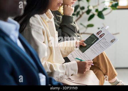 Junge Erwachsene multiethnische Gruppe, die im Wartebereich des Visumzentrums sitzt, junge Erwachsene Frau mit Reisepass und Visumantragsformular mit Stift, Vorbereitung der Dokumente für die Verarbeitung Stockfoto
