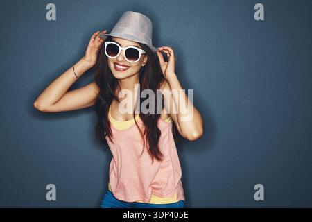 Mode, Porträt und Frau mit Sonnenbrille im Studio für lässiges, trendiges oder cooles Outfit auf Platz. Lächeln, Hut und weibliche Person mit Selbstvertrauen Stockfoto